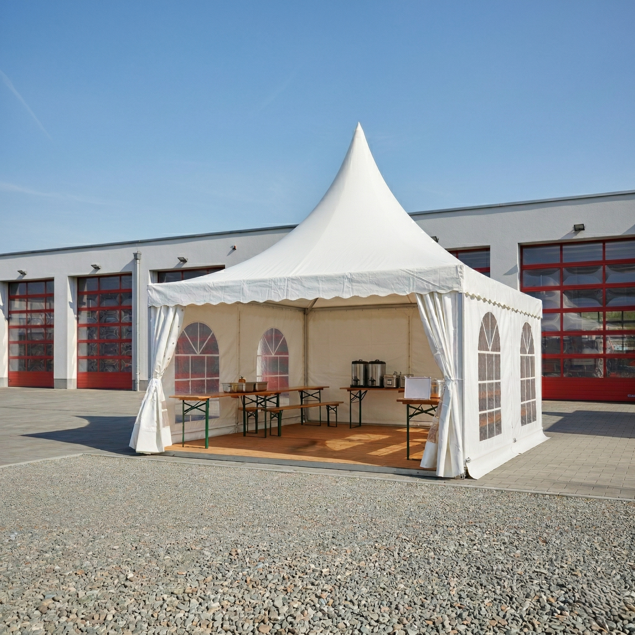 Ein quadratisches Produktbild eines weißen Pagodenzelts, das auf einer gepflasterten Fläche vor einem modernen Gebäude (Feuerwehrwache) mit großen roten Rolltoren steht. Das Zelt hat ein charakteristisches, hohes Spitzdach und ist an zwei Seiten offen, wodurch der Blick auf den Innenraum mit einem hellen Holzboden und einfachen Holztischen frei wird. Die Szenerie ist menschenleer, wirkt ultrarealistisch und wird von klarem, natürlichem Tageslicht unter blauem Himmel beleuchtet.